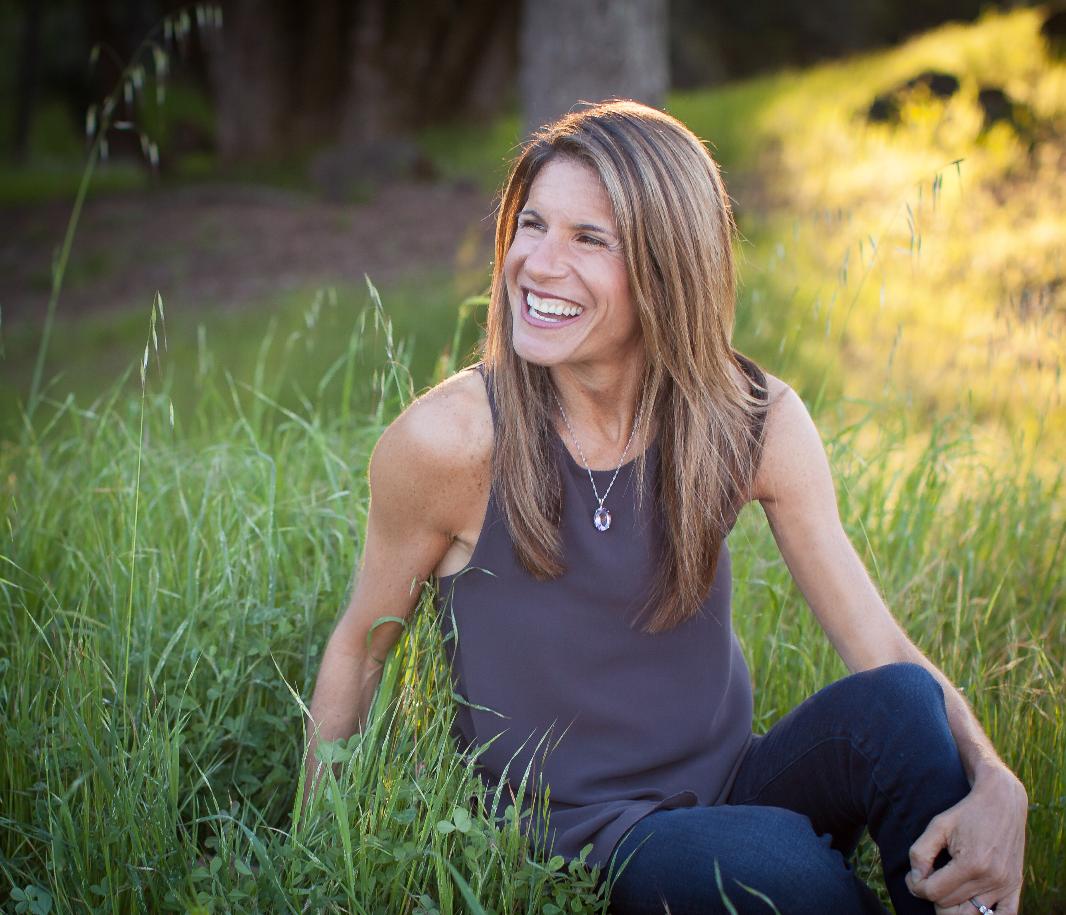 Sheryl O'Loughlin, entrepreneur, CEO, seated in grass with sun, smiling. looking off to the side