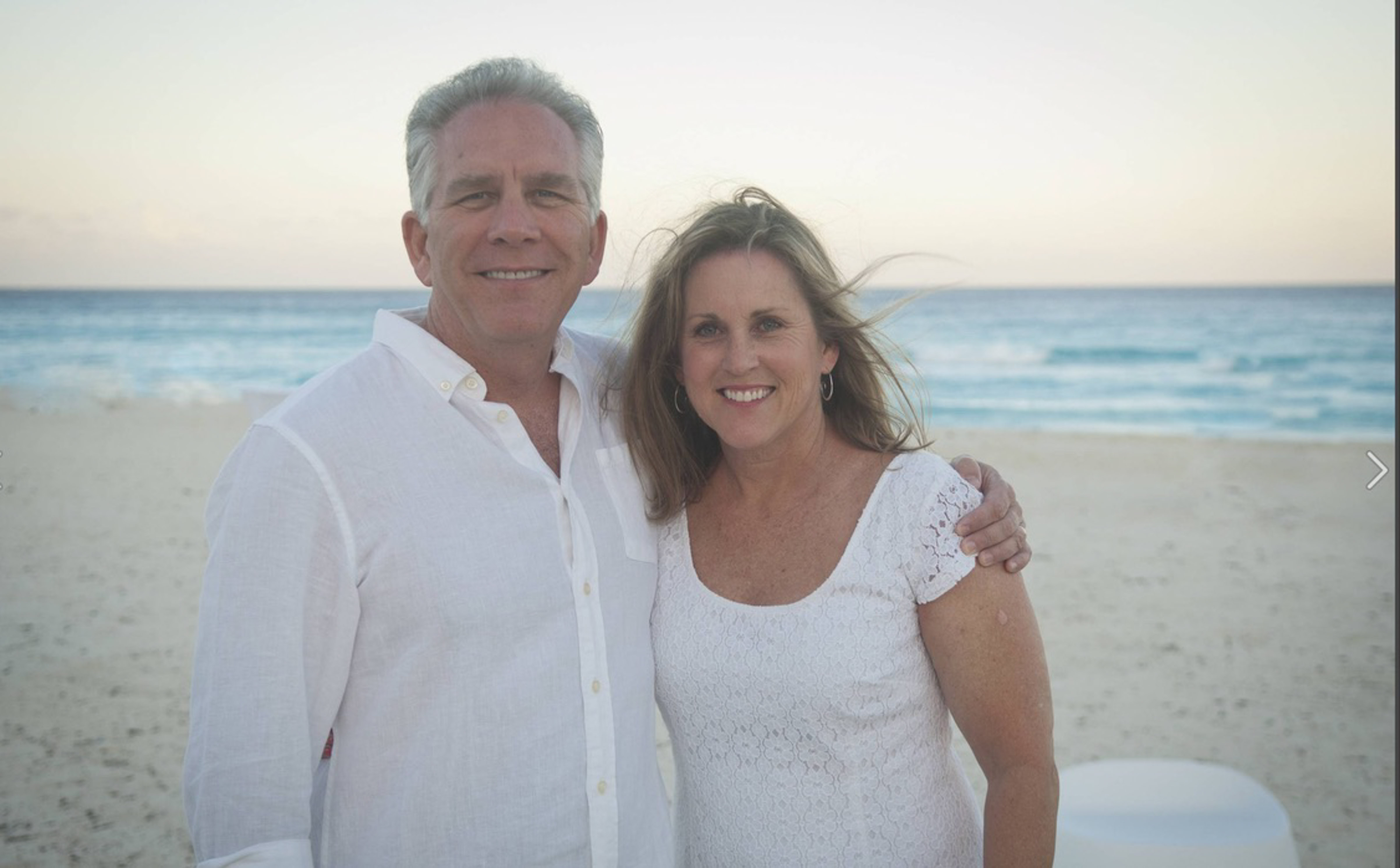 middle aged couple dressed in white, standing on the beach