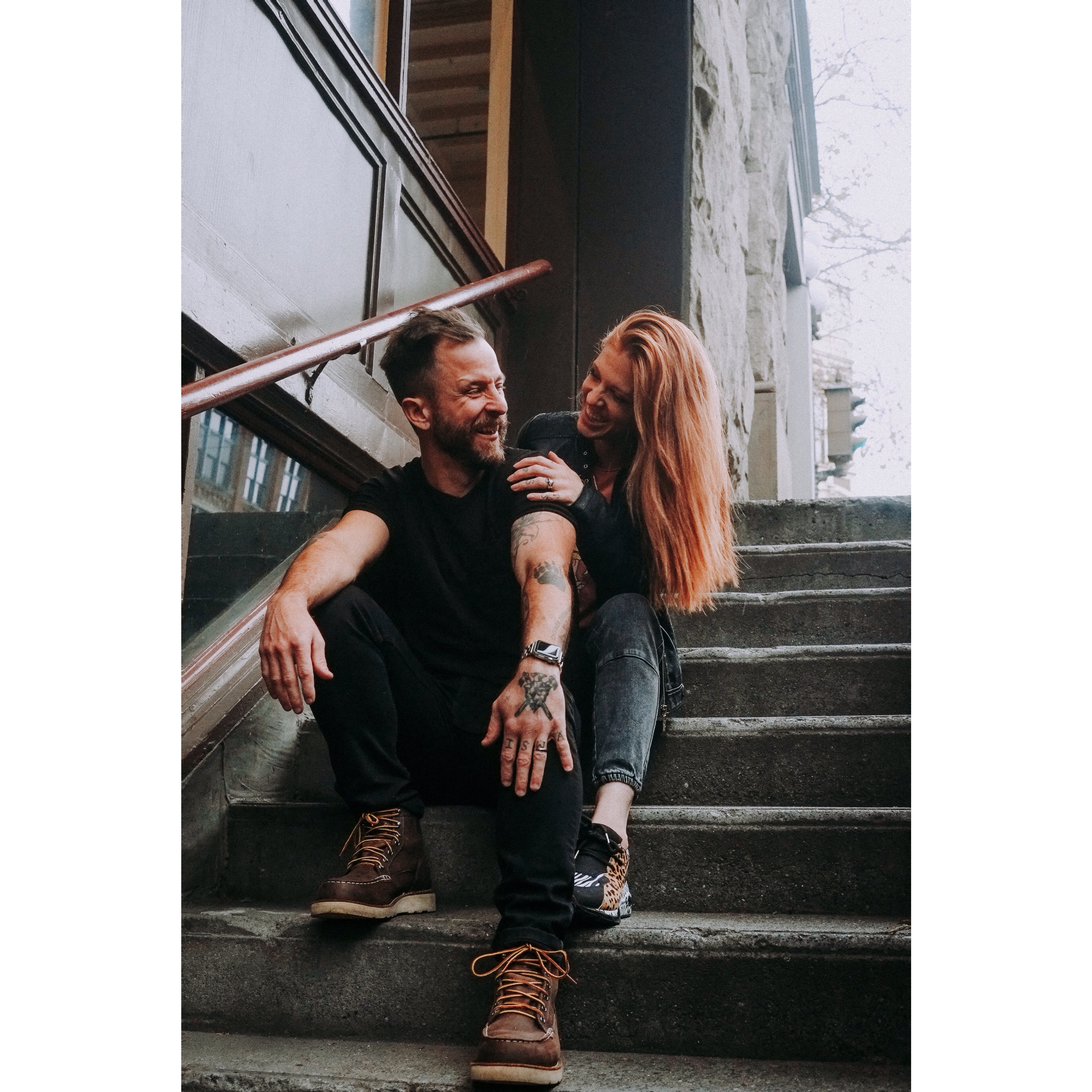 Young couple with tattoos sitting on steps, she's behind him, smiling at each other